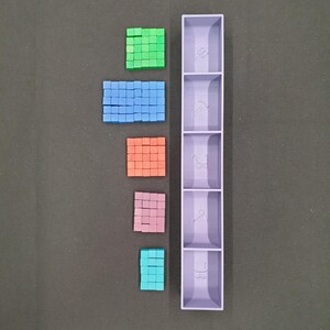 May include: A top-down view displays an assortment of small, square blocks in green, blue, orange, pink, and teal. A tall, lavender organiser with five compartments is to the right of the blocks. The background is a dark grey surface.