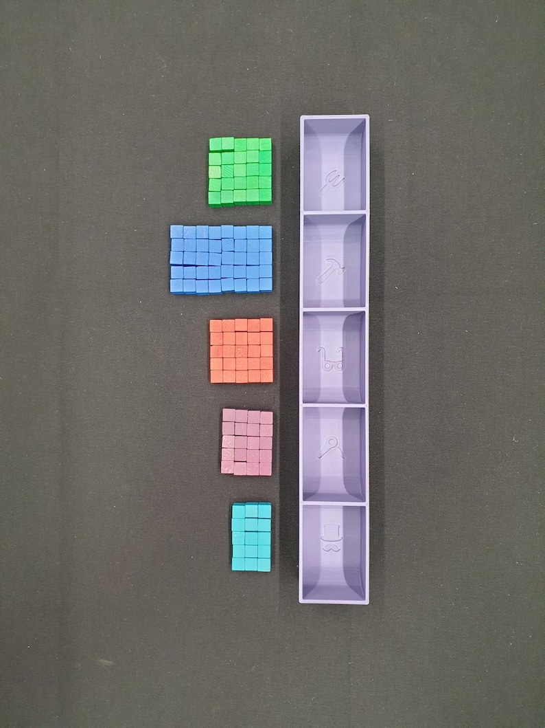 May include: A top-down view displays an assortment of small, square blocks in green, blue, orange, pink, and teal. A tall, lavender organiser with five compartments is to the right of the blocks. The background is a dark grey surface.