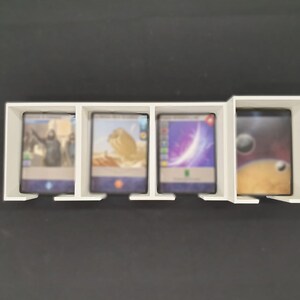 May include: A white card holder with four sections, each containing a trading card. The cards display colourful images, including a person, desert scene, and space. The holder is on a black surface.