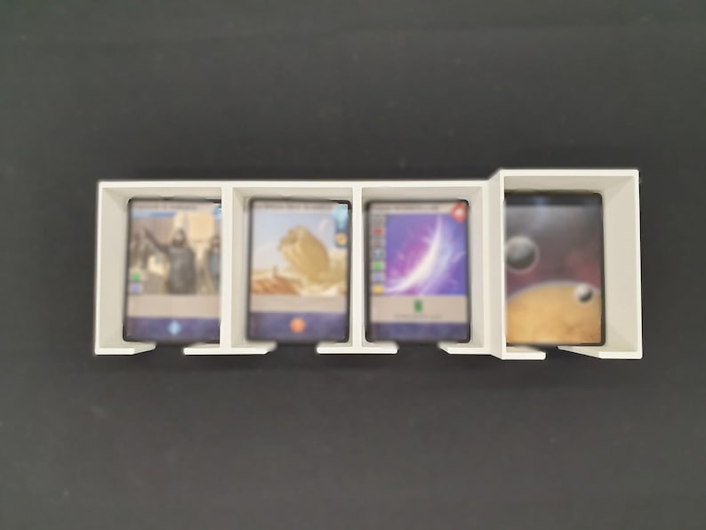 May include: A white card holder with four sections, each containing a trading card. The cards display colourful images, including a person, desert scene, and space. The holder is on a black surface.