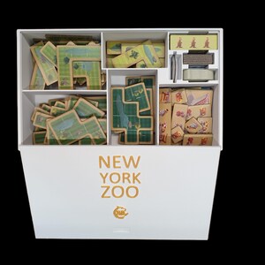 May include: A white box with the words "NEW YORK ZOO" printed on the front. The box is open and contains various game pieces, including tiles with green and brown designs, and small wooden tiles with colorful designs.