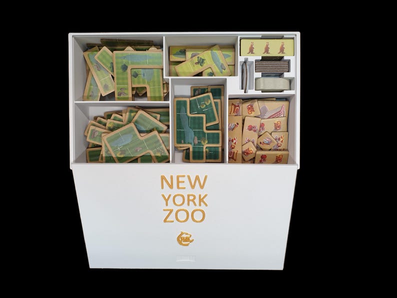 May include: A white box with the words "NEW YORK ZOO" printed on the front. The box is open and contains various game pieces, including tiles with green and brown designs, and small wooden tiles with colorful designs.