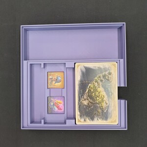May include: A lilac-coloured game box containing a deck of cards and two small game pieces. The cards display a landscape image. The box is open, showing the contents, against a dark background.