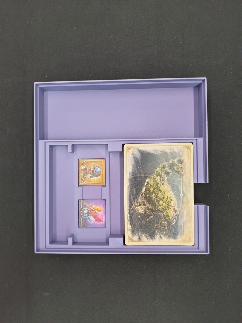 May include: A lilac-coloured game box containing a deck of cards and two small game pieces. The cards display a landscape image. The box is open, showing the contents, against a dark background.
