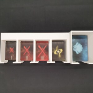 May include: A white plastic card holder with five compartments. Four compartments hold cards with red and gold graphics. The fifth compartment holds a card with a blue and white design. The holder sits on a black surface.