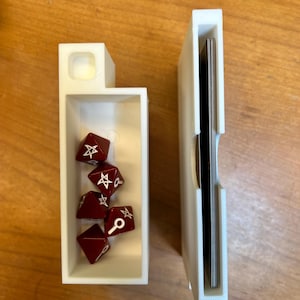 May include: Two white 3D-printed game component holders. One contains several dark red polyhedral dice with white symbols. The other holds a stack of black-edged cards. The holders are on a light brown wooden surface.
