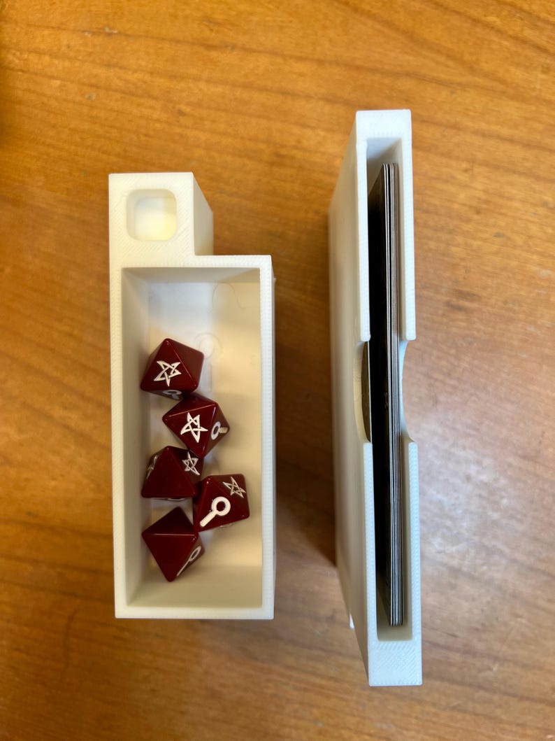 May include: Two white 3D-printed game component holders. One contains several dark red polyhedral dice with white symbols. The other holds a stack of black-edged cards. The holders are on a light brown wooden surface.