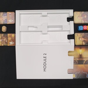May include: A board game set with cards, dice, and a white box labeled "MODULE 2". The cards feature illustrations of people and symbols. Three dice are blue, red, and yellow. A wooden token is also present.