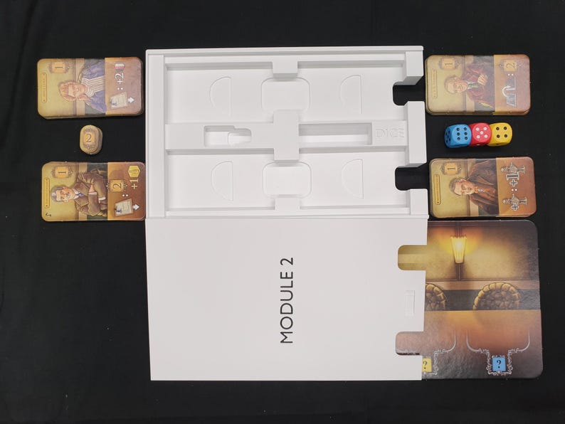 May include: A board game set with cards, dice, and a white box labeled "MODULE 2". The cards feature illustrations of people and symbols. Three dice are blue, red, and yellow. A wooden token is also present.