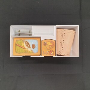Puede incluir: Un organizador de cart&oacute;n blanco lleno de componentes de juego. El contenido incluye cartas ilustradas, una funda de cuero sint&eacute;tico color beige y otras piezas peque&ntilde;as. Las cartas presentan dibujos de alimentos y otros objetos. Las piezas del juego est&aacute;n dispuestas.