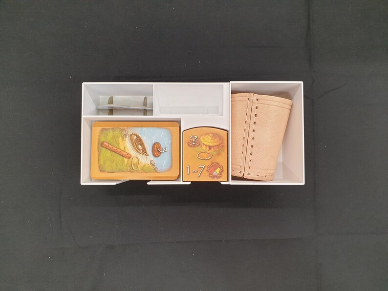 Puede incluir: Un organizador de cart&oacute;n blanco lleno de componentes de juego. El contenido incluye cartas ilustradas, una funda de cuero sint&eacute;tico color beige y otras piezas peque&ntilde;as. Las cartas presentan dibujos de alimentos y otros objetos. Las piezas del juego est&aacute;n dispuestas.