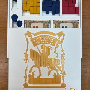 Op de afbeelding: Een witte speldoos met een gouden uitsnede van een ridder te paard. De tekst "The King is Dead" staat op een banner boven de ridder. De doos heeft drie compartimenten met stapels rode, blauwe en gele blokken.