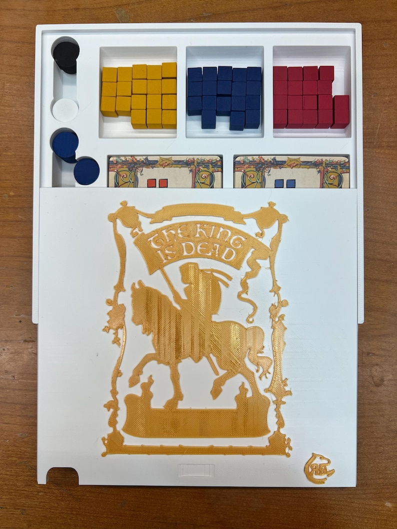 Op de afbeelding: Een witte speldoos met een gouden uitsnede van een ridder te paard. De tekst "The King is Dead" staat op een banner boven de ridder. De doos heeft drie compartimenten met stapels rode, blauwe en gele blokken.