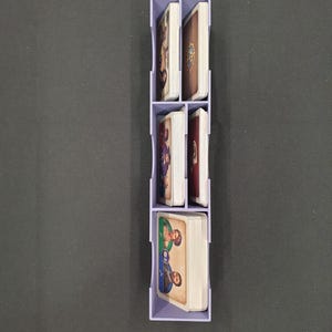 May include: A light purple card organiser, vertically arranged, filled with stacks of illustrated cards. The organiser has four compartments, each holding a deck of cards with colourful artwork. The cards appear to be for a board game.