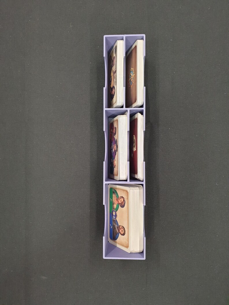 May include: A light purple card organiser, vertically arranged, filled with stacks of illustrated cards. The organiser has four compartments, each holding a deck of cards with colourful artwork. The cards appear to be for a board game.