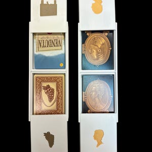 May include: Two white boxes with lids, each containing three cards. The top card in the left box is blue with the word "VENDUTA" printed on it. The middle card in the left box is brown with a bunch of grapes on it. The top card in the right box is blue with a gold oval portrait of a woman. The middle card in the right box is blue with a gold oval portrait of a woman.