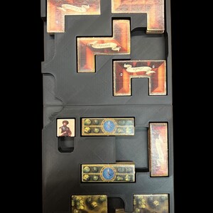 May include: A black plastic storage box with compartments for organizing game pieces. The box contains a variety of game pieces, including wooden tiles, cards, and a small figure.