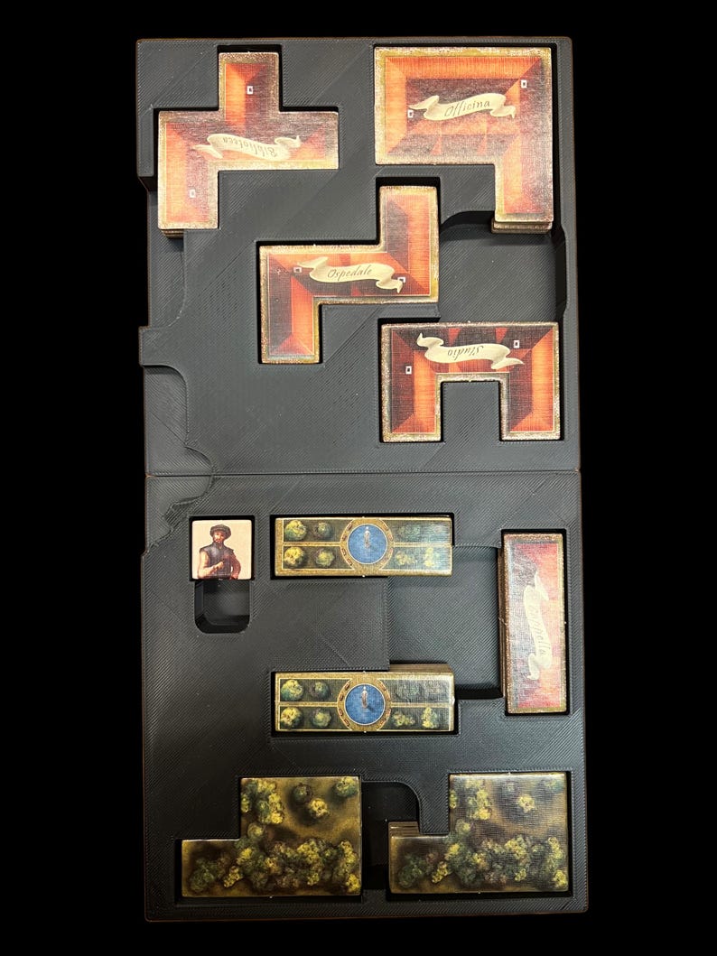 May include: A black plastic storage box with compartments for organizing game pieces. The box contains a variety of game pieces, including wooden tiles, cards, and a small figure.