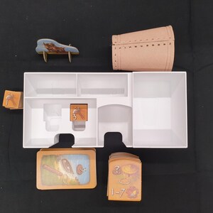 Puede incluir: Un organizador de juegos de mesa con varios componentes. Incluye una caja blanca con compartimentos, un barril en miniatura, un peque&ntilde;o banco de madera, cartas con ilustraciones y fichas numeradas. Las piezas del juego est&aacute;n dispuestas sobre una superficie oscura.