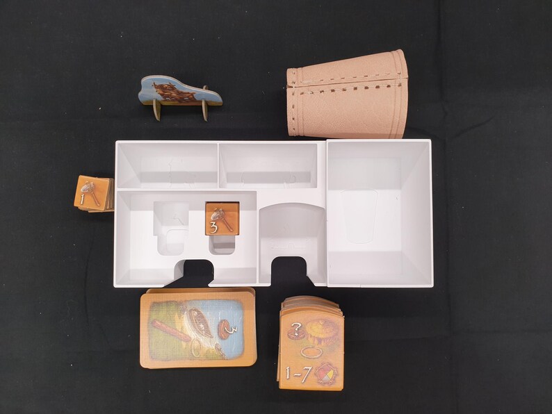 Puede incluir: Un organizador de juegos de mesa con varios componentes. Incluye una caja blanca con compartimentos, un barril en miniatura, un peque&ntilde;o banco de madera, cartas con ilustraciones y fichas numeradas. Las piezas del juego est&aacute;n dispuestas sobre una superficie oscura.