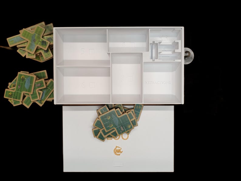 May include: White box with compartments for storing game pieces. The box is open and the compartments are labeled "SET", "ACTION", and "SET". There are stacks of green and brown game pieces outside of the box.