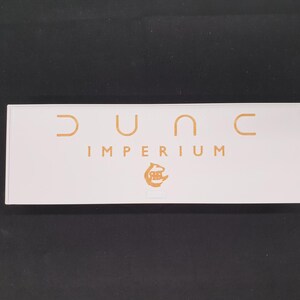 May include: A rectangular white box with the words "DUNE IMPERIUM" in gold lettering. The box also features a gold logo. The background is a solid black colour.