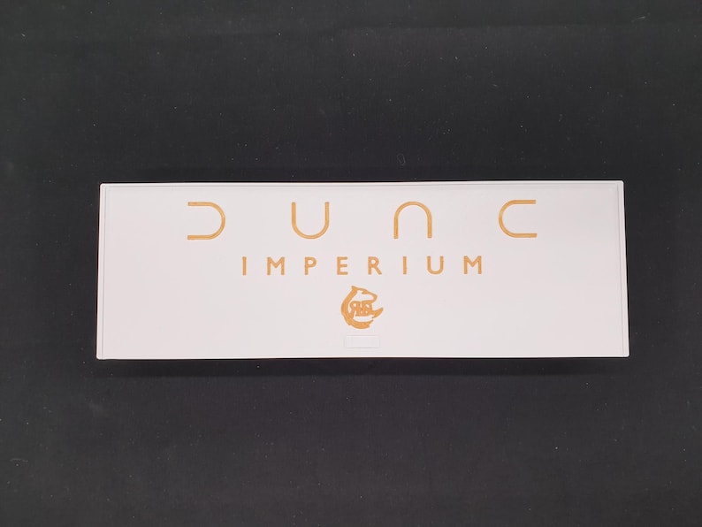 May include: A rectangular white box with the words "DUNE IMPERIUM" in gold lettering. The box also features a gold logo. The background is a solid black colour.