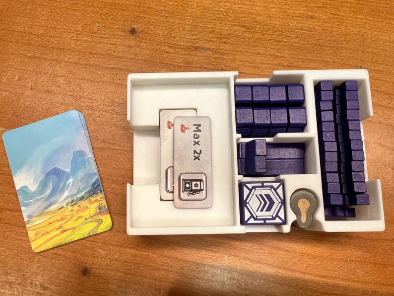 K&ouml;nnte beinhalten: Ein wei&szlig;er Plastik-Organizer mit F&auml;chern zur Aufbewahrung von Spielsteinen. Der Organizer enth&auml;lt zwei Karten mit dem Text "Max 2x" und einem Symbol f&uuml;r eine Zapfs&auml;ule. Au&szlig;erdem gibt es blaue Plastikbl&ouml;cke und eine blaue Plastikfliese mit einem Pfeilsymbol.