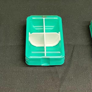 以下が含まれることがあります： 3つの長方形のターコイズ色のプラスチック容器。1つは空で、2つ目は白いインサートと白い紐があり、3つ目は白いインサートがあります。容器は積み重ねられており、リブのデザインになっています。