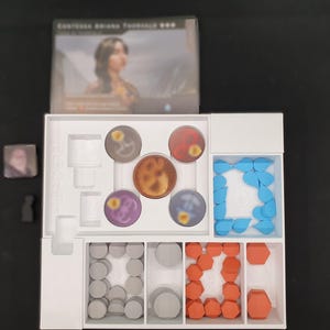 May include: A board game set with a white interior organiser. The set includes circular game pieces in various colours, including brown, red, purple, and blue. There are also grey and orange geometric shapes. The game box features a woman's portrait.