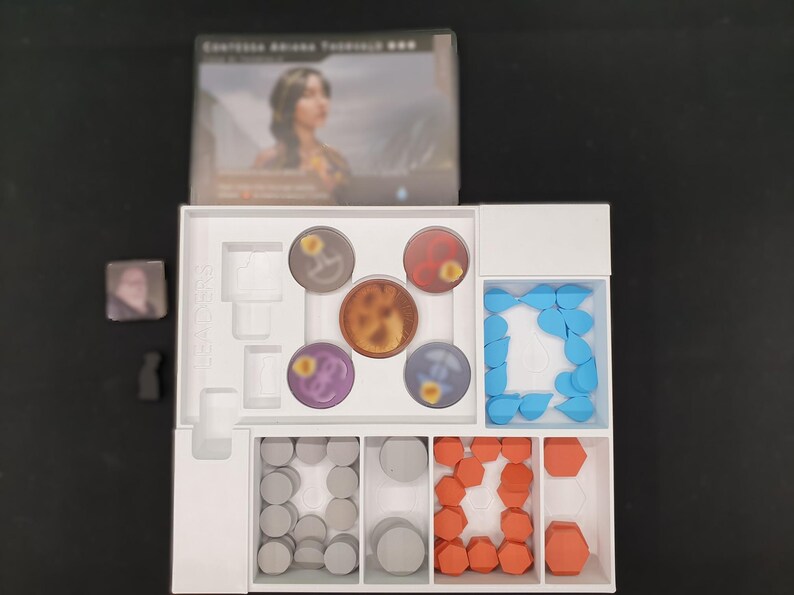 May include: A board game set with a white interior organiser. The set includes circular game pieces in various colours, including brown, red, purple, and blue. There are also grey and orange geometric shapes. The game box features a woman's portrait.