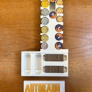 Pode incluir: Organizador de pl&aacute;stico branco para o jogo de tabuleiro Autobahn. Tem ranhuras para diferentes pe&ccedil;as de jogo coloridas e um compartimento para pe&ccedil;as de madeira. A tampa tem o logotipo do jogo, "AUTOBAHN", em letras douradas.