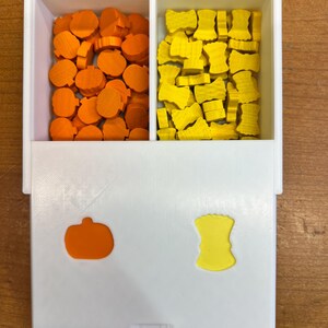 May include: A white box with two compartments. The left compartment is filled with orange pumpkin-shaped game pieces. The right compartment is filled with yellow bow-shaped game pieces. The box has a small orange pumpkin and a yellow bow on the front.