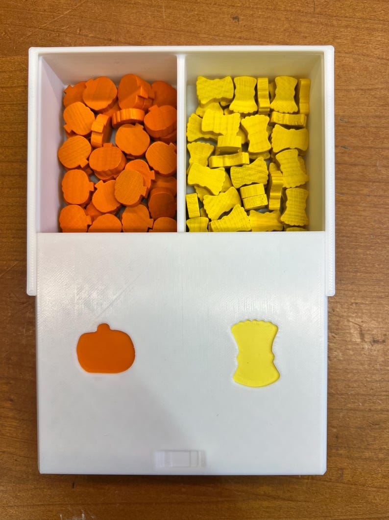 May include: A white box with two compartments. The left compartment is filled with orange pumpkin-shaped game pieces. The right compartment is filled with yellow bow-shaped game pieces. The box has a small orange pumpkin and a yellow bow on the front.