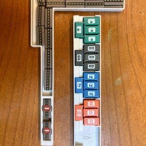 K&ouml;nnte beinhalten: Ein wei&szlig;es Kunststofftablett mit einer grauen Bahn und einer Vielzahl von farbigen Beh&auml;ltern. Die Beh&auml;lter sind in Reihen gestapelt und mit verschiedenen Symbolen gekennzeichnet. Das Tablett ist f&uuml;r ein Brettspiel konzipiert.