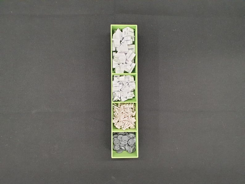 May include: A green plastic organizer with five compartments, each filled with small, gray components. The components vary in shape, including rectangular blocks, irregular shapes, and small, round pieces. The organizer sits on a dark gray surface.