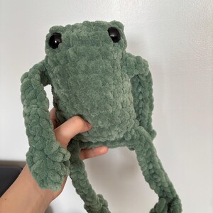 May include: A handmade, plush frog in a soft, sage green color. The frog has large, black button eyes and outstretched limbs. The textured yarn gives the frog a cuddly appearance, perfect for a child's toy or room decor.