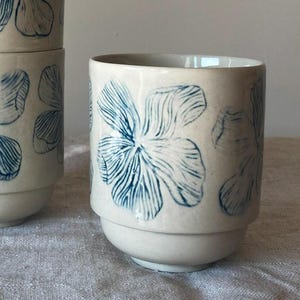 Op de afbeelding: Drie witte keramische bekers met blauwe bloemenmotieven. De bekers zijn op elkaar gestapeld.
