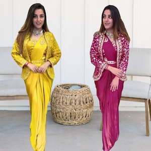 May include: Two women wearing matching embroidered jackets over yellow and pink draped sarees with matching belts. The women are standing in front of a white wall and a wicker basket.