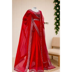 Peut inclure: Un sari rouge vif pr&eacute;sent&eacute; sur un mannequin, orn&eacute; de broderies argent&eacute;es complexes le long des bordures et du corsage. Le tissu transparent est agr&eacute;ment&eacute; de petites d&eacute;corations &eacute;parses et une ceinture assortie marque la taille.