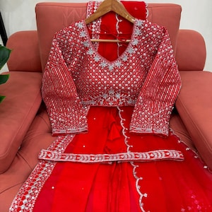 Peut inclure: Un ensemble compos&eacute; d'un chemisier rouge &agrave; manches longues et d'une jupe assortie, orn&eacute; de broderies argent&eacute;es et de sequins. Le chemisier pr&eacute;sente un d&eacute;collet&eacute; en V et des motifs g&eacute;om&eacute;triques, avec une ceinture d&eacute;corative argent&eacute;e.
