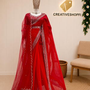 Peut inclure: Un sari rouge vif en tissu filet transparent avec des broderies argent&eacute;es complexes et des d&eacute;tails en miroir le long des bordures. Le v&ecirc;tement comprend un chemisier assorti avec des ornements d&eacute;taill&eacute;s et une ceinture, pr&eacute;sent&eacute; sur un mannequin.