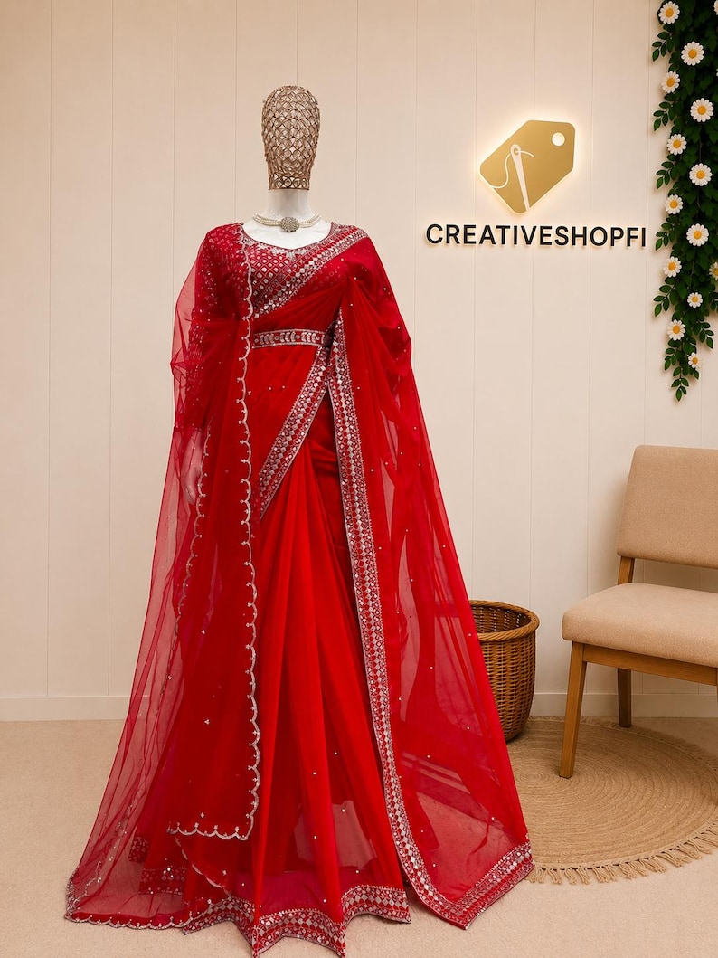 Peut inclure: Un sari rouge vif en tissu filet transparent avec des broderies argent&eacute;es complexes et des d&eacute;tails en miroir le long des bordures. Le v&ecirc;tement comprend un chemisier assorti avec des ornements d&eacute;taill&eacute;s et une ceinture, pr&eacute;sent&eacute; sur un mannequin.
