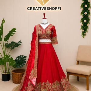 May include: A vibrant red lehenga set featuring a cropped blouse with a V-neckline and three-quarter sleeves, paired with a voluminous floor-length skirt. Both pieces are adorned with intricate gold embroidery and a matching red dupatta.