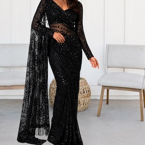 May include: A black saree featuring intricate lace embroidery and sequin embellishments throughout the fabric. The garment includes long sleeves, a V-neckline, and a sheer midriff section, with a long, flowing pallu draped over one shoulder.
