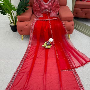 Peut inclure: Un ensemble lehenga rouge vif compos&eacute; d'un chemisier &agrave; manches longues avec des broderies argent&eacute;es complexes et des miroirs. Le dupatta et la jupe en voile rouge assortis sont orn&eacute;s de bordures argent&eacute;es, de bords festonn&eacute;s et de perles.
