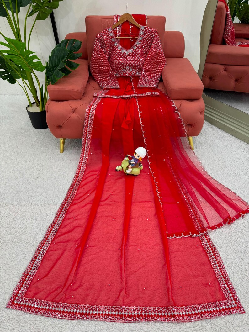 Peut inclure: Un ensemble lehenga rouge vif compos&eacute; d'un chemisier &agrave; manches longues avec des broderies argent&eacute;es complexes et des miroirs. Le dupatta et la jupe en voile rouge assortis sont orn&eacute;s de bordures argent&eacute;es, de bords festonn&eacute;s et de perles.