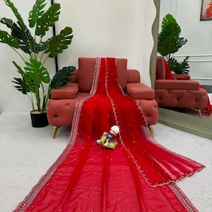 Peut inclure: Un drap&eacute; en tissu rouge vif et transparent, orn&eacute; de broderies argent&eacute;es complexes sur les bordures et de petites d&eacute;corations blanches &eacute;parpill&eacute;es. Le textile est pr&eacute;sent&eacute; &eacute;tal&eacute; sur une surface, agr&eacute;ment&eacute; d'une petite figurine.
