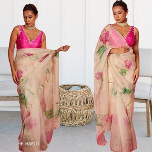 Puede incluir: Un sari de gasa de color melocotón con un estampado floral de flores de loto rosas y hojas verdes. El sari tiene un ribete dorado y se combina con una blusa rosa fucsia sin mangas.