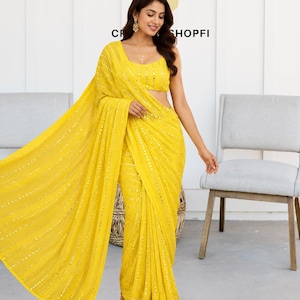 Puede incluir: Un sari amarillo brillante con intrincados detalles de lentejuelas. El sari está elegantemente drapeado, mostrando su tejido fluido y diseño. También se muestra un top a juego. El texto "SHOPFI" es visible.
